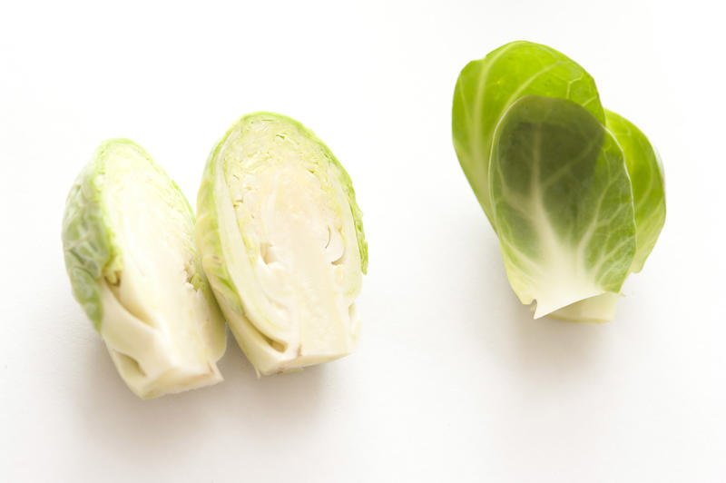 Halved fresh uncooked Brussel sprout with loose green outer leaves alongside over a white background
