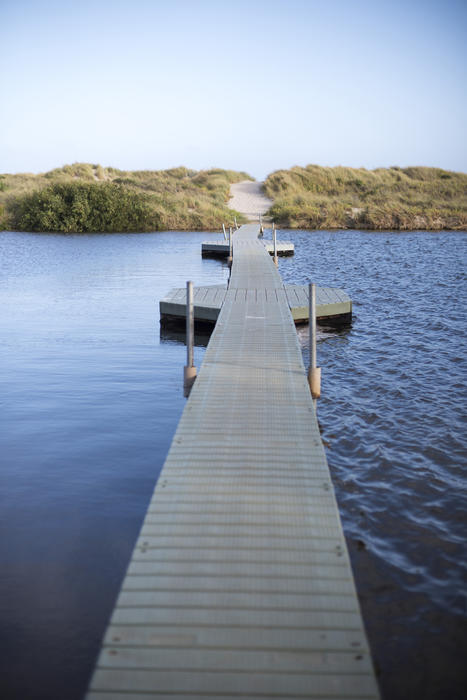 Wooden floating pedestrian bridge over water with calm sheltered sea on one side and rippling rough water the other in a conceptual image