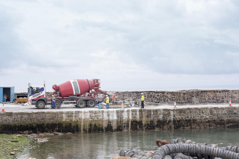 Red cement mixer truck park on a wharf or quay on a construction site with workmen in hardhats under a cloudy sky