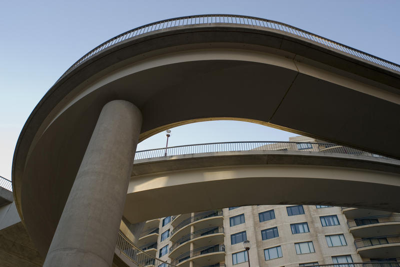 Looping concrete walkway viewed from below against blue sky with urban high-rise buildings in the background