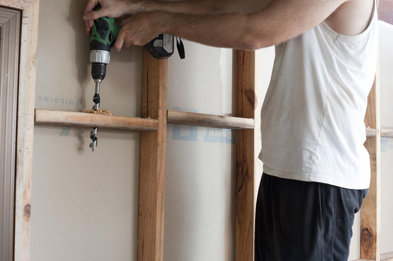 Man in white shirt drilling cable hole in the stud wall during frame house construction process