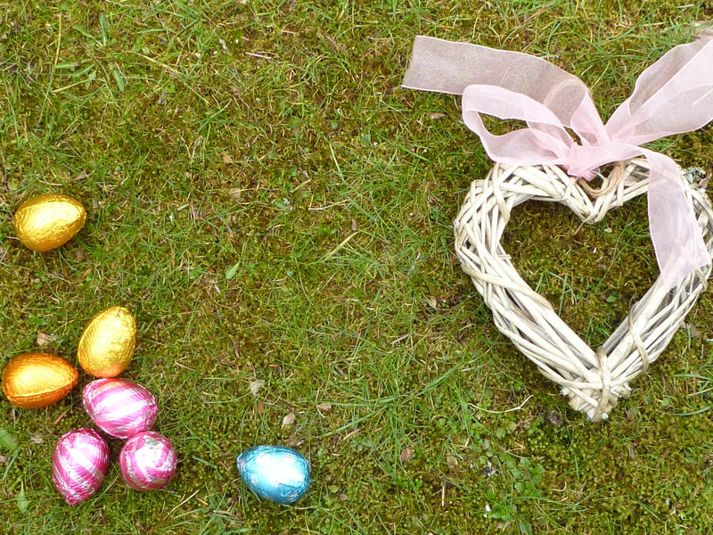 Easter egg hunt and love concept. Colorful chocolate eggs in foil and wicker heart with pink ribbon bow viewed from above on the grass with copy space