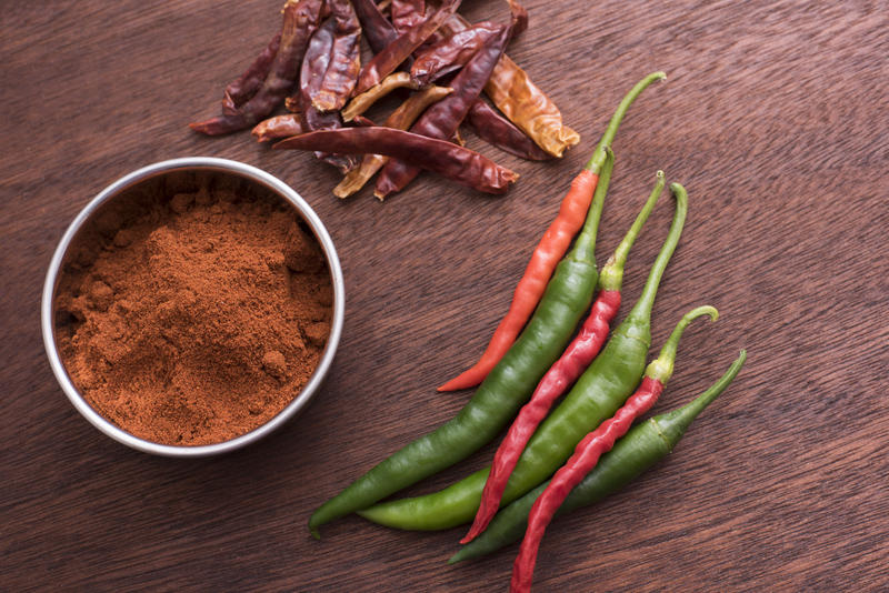 Fresh green and red, pile of dried and a bowl of powdered chilli pepper on brown wooden kitchen table viewed from above