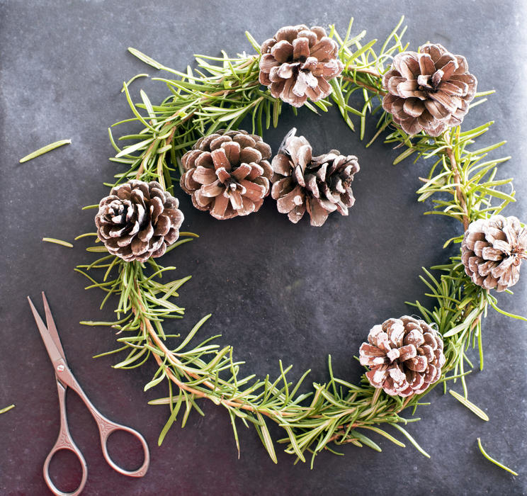 Homemade Christmas wreath with pine foliage and cones alongside a pair of scissors over a textured dark background