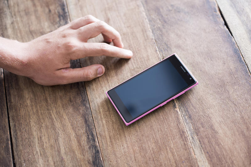 Male hand alongside a blank cellphone or smartphone on a wooden table with the black screen visible to the viewer