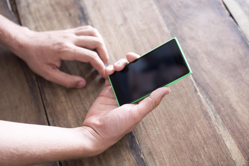 A close up of a man holding a blank, black smartphone in his hand on a timber bench top background.