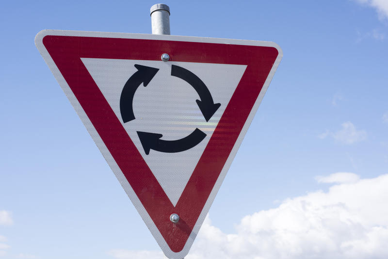 Traffic warning sign for an approaching roundabout or circle against a cloudy blue sky