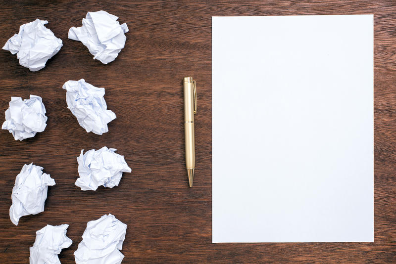 Trouble getting started concept with balls of crumpled paper alongside a blank white sheet with pen on a wooden desk signifying writers block or lack of ideas and inspiration