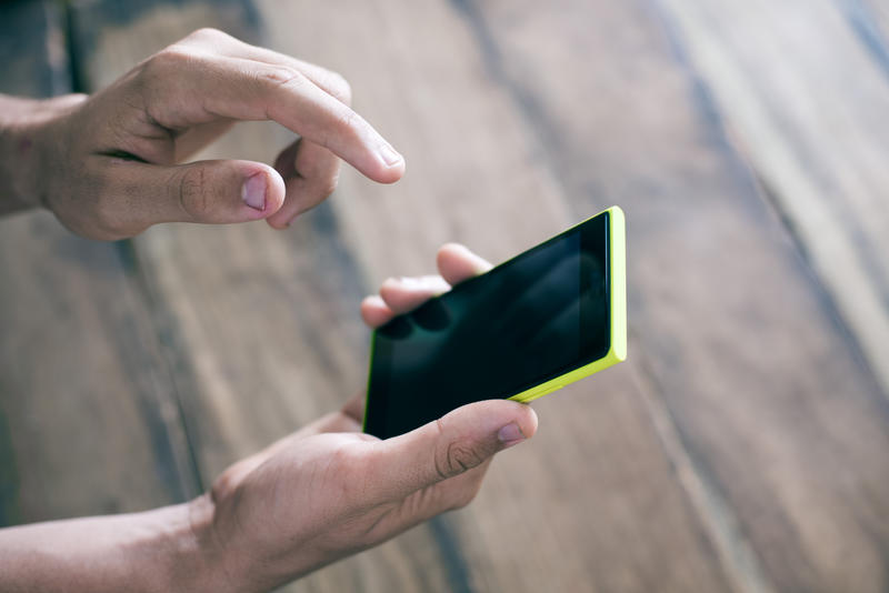 A close up of a man holding and using a smartphone with a blank, black screen against a plain blurred background.