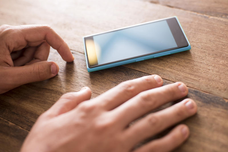 Man waiting for contact on his mobile phone resting his hands on a wooden table on either side of the blank cellphone in a close up view