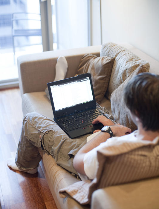 Businessman remote working from home reclining on a sofa with his laptop in a Covid-19 concept viewed from the rear