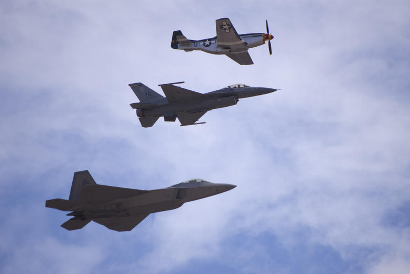 three planes flying past, a P-51 mustang with propeller engine, and modern FA18 and F22 jet aircraft