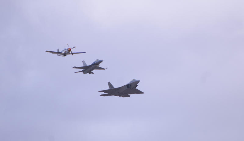 three planes in flight, a P-51 mustang with propeller, and FA18 and F22 jet aircraft