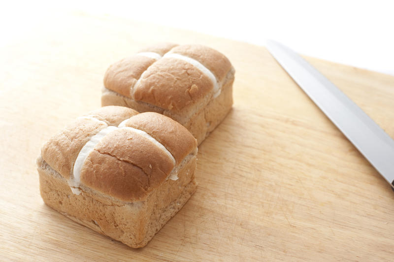 Two fresh square traditional Hot Cross Buns with frosted crosses on a wooden board with a knife.