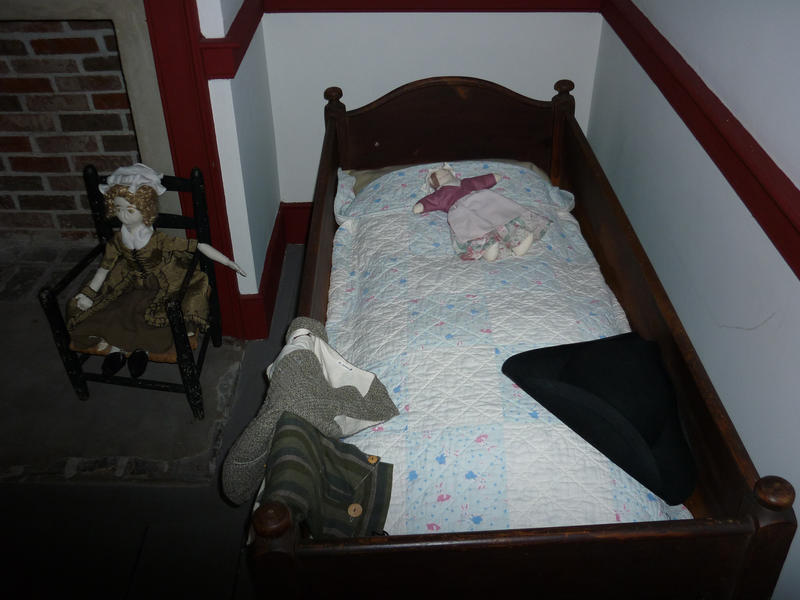 Small antique wooden childs bed set in an alcove with a vintage doll sitting in a small chair alongside it