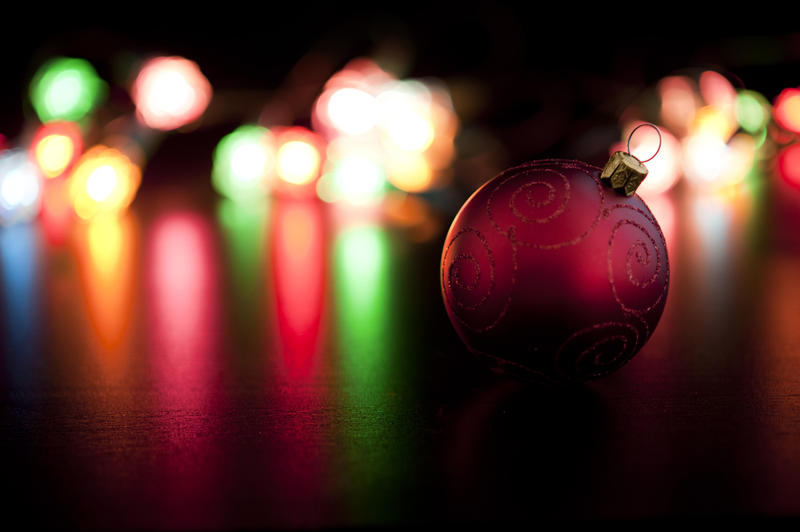 A single red bauble decoration lies in front of a background of colourful Christmas lights shining across a reflective surface
