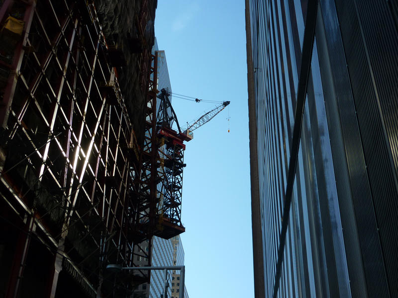 View looking upwards between highrise buildings on a narrow street to a crane suspended from a building facade that is still under constrcution and covered in scaffolding