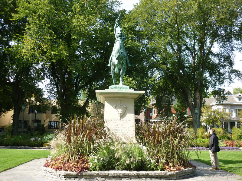Frontal view of a Joan of Arc statue with Joan astride a horse on a plinth standing in a small flowerbed in a park with a man standing admiring it