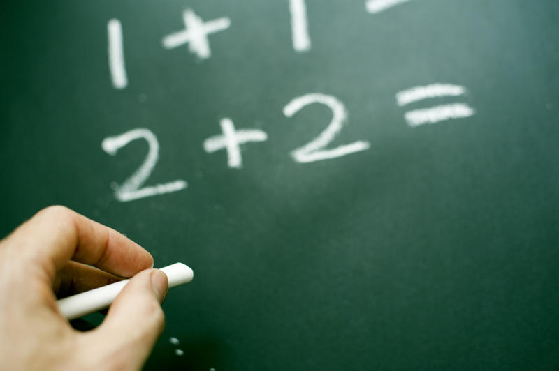 Teachers hand doing mathematics writing basic sums on a blackboard with chalk to teach kindergarten children addition and simple mathematics