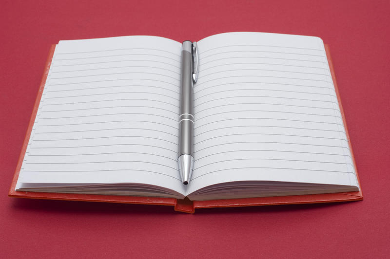 Open blank lined notebook with a ballpoint pen lying down the centre on a red studio background ready for your message or text