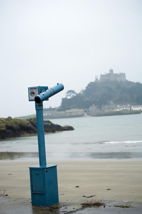 A view from the mainland beach of a tourist viewing telescope pointing towards the offshore tidal island of St Michaels Mount with its historical castle, village and monastery in the mist