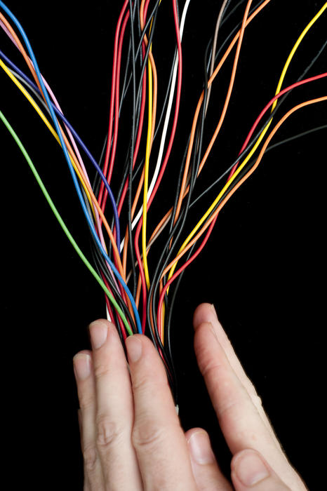 Splayed multiple brightly coloured electric wires converging between two human hands on a black background