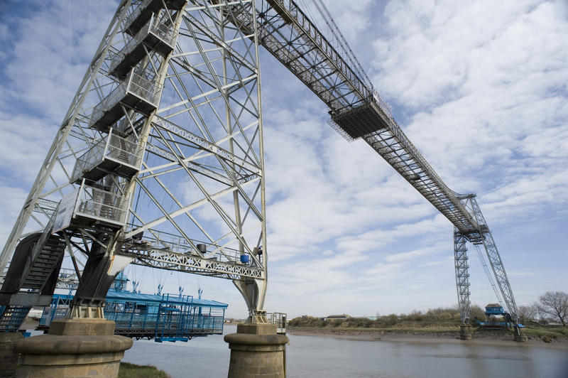 The blue gondola suspended close to the bank below the metal span of the historical Newport Transporter Bridge ferrying cars and people acros the River Usk