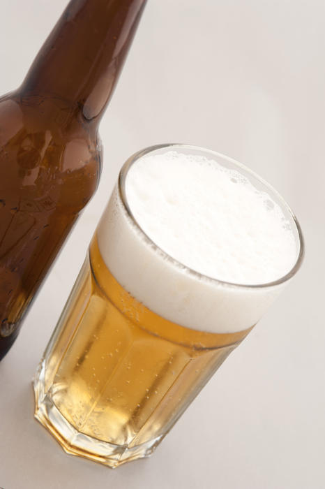 Glass of beer with a frothy head alongside a brown unlabeled beer bottle, tilted high angle view on white