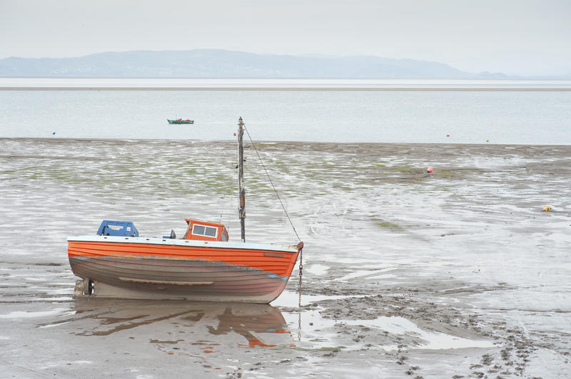 Small colourful orange wooden fishing boat at low tide left high and dry on the sand by the receding water at Morecambe