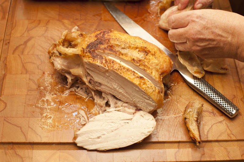Partially consumed roast chicken with a slice of the breast lying alongside and a mans hands visible at the top of the frame, view from above on a wooden chopping board
