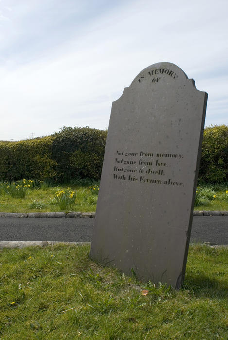 Blank headstone with poetic inscription standing at an angle alongside a small rural lane in open countryside