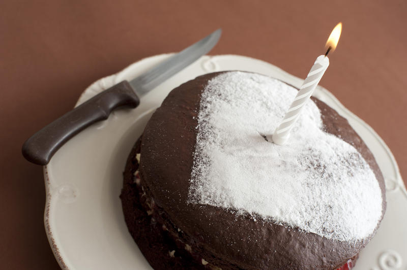 Close up Lighted Candle on Chocolate Cake, Decorated with Heart Shape Sugar on Top, Placed on a Plate with Knife on the Side.