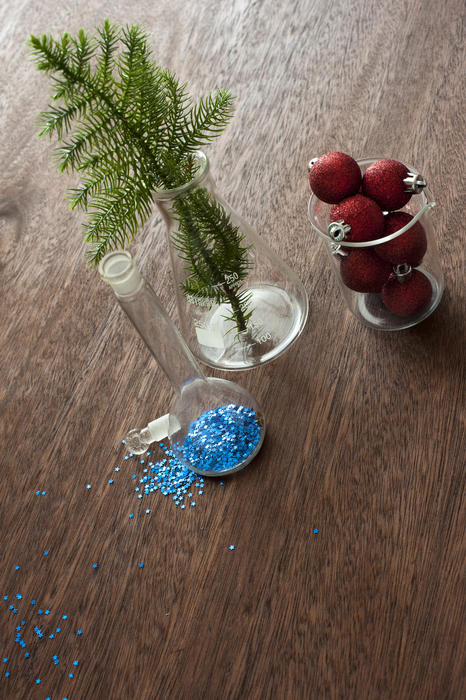 High Angle View Christmas Chemisty Concept Still Life - Small Evergreen Branch, Blue Glitter and Red Ornaments in Various Sized Glass Science Beakers on Textured Wooden Table with Copy Space