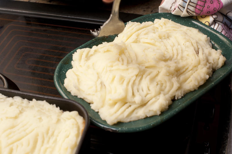 Person preparing cottage pie with mashed potato topping on minced meat making a pattern on it with a fork before baking it in the oven