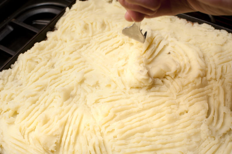 Cottage pie preparation with a cook smoothing the mashed potato crust over the meat with a fork before baking or grilling
