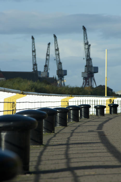 Large industrial cranes in a dockyard for shipbuilding or offloading cargo with a row of black bollards in the foreground curving off to the right