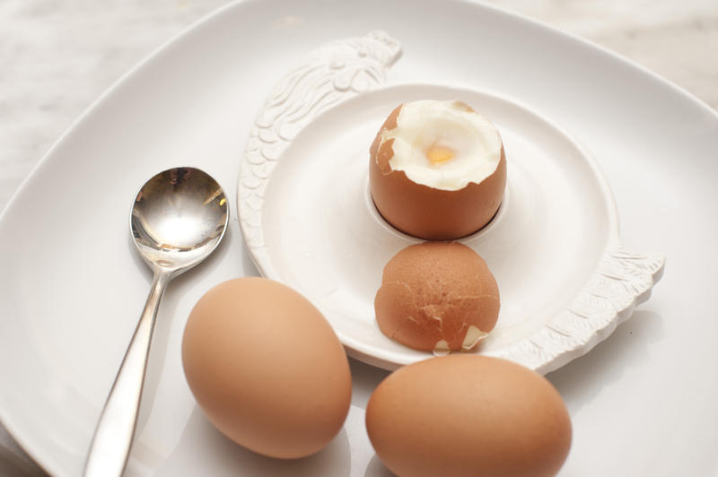 Serving of three boiled eggs for breakfast with two eggs on the side and the third soft-boiled egg with its top cut off in an eggcup