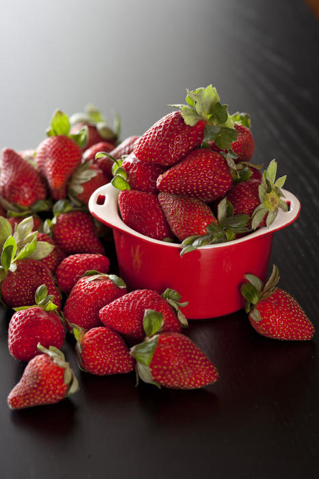 Ripe red strawberries in a small ramekin overflowing onto a dark surface with a highlight behind for copyspace