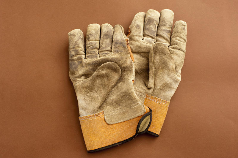 Pair of old leather gardening gloves with creased fingers and stains lying on a brown background