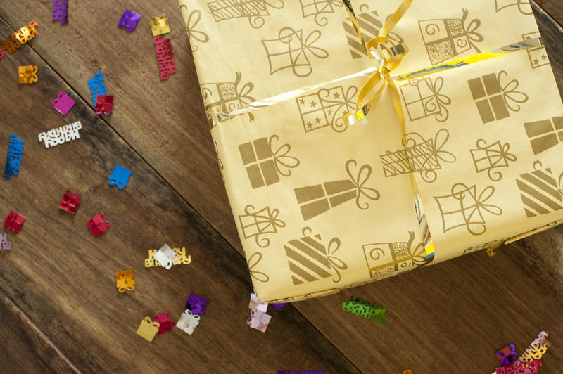 Gift-wrapped birthday present in pretty cream paper patterned with gifts, overhead view on a wooden table with colorful Happy Birthday sprinkles