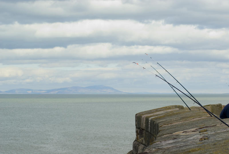 Row of fishing rods protruding over a stone sea wall at Whitehaven as fishermen enjoy some recreational angling
