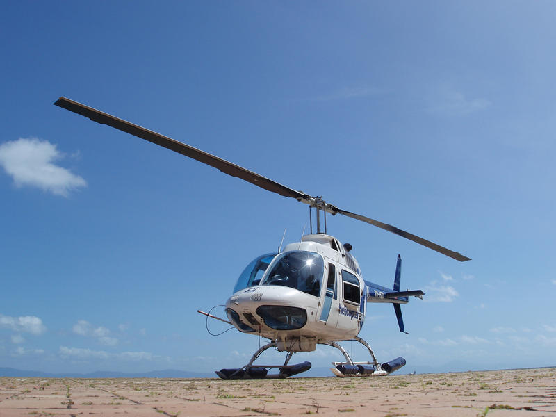 Helicopter parked on a helipad against a sunny blue sky with the rotor blade forming a diagonal line through the frame