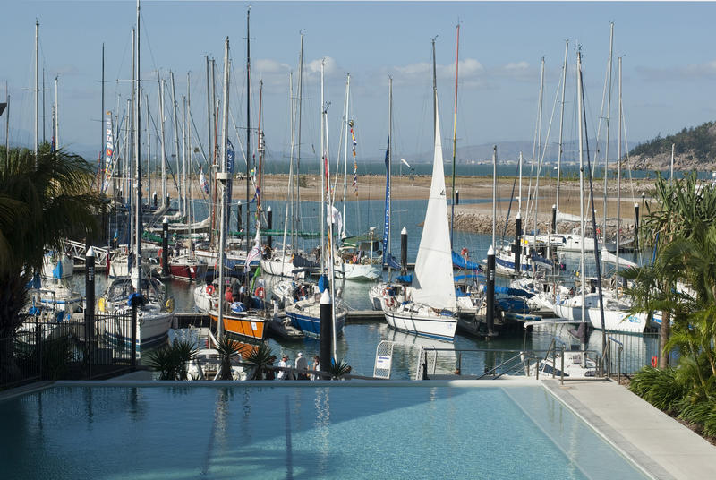Small marina in a holiday resort with moored yachts and sail boats viewed across the resort swimming pool