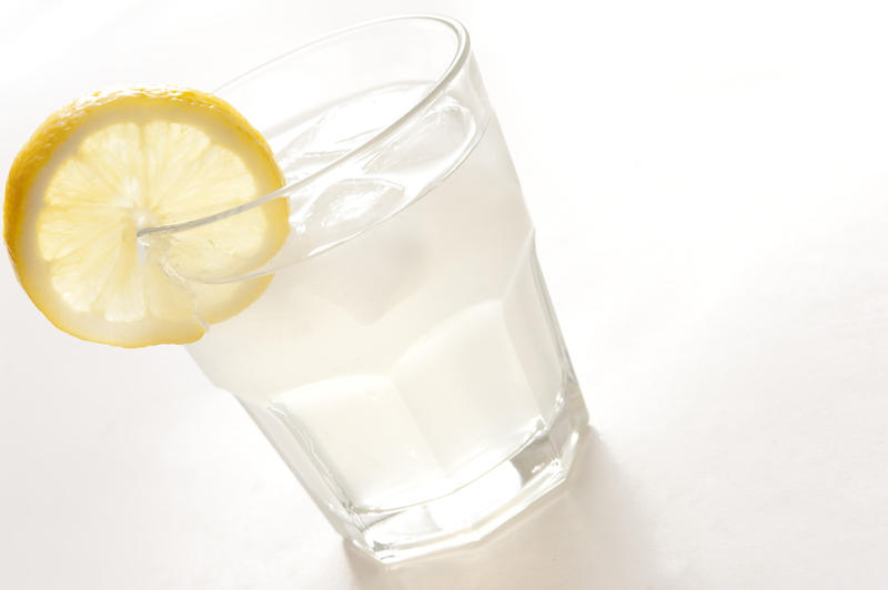 Long glass of clear spirits served with ice and garnished with a slice of fresh lemon, tilted angle close up on white