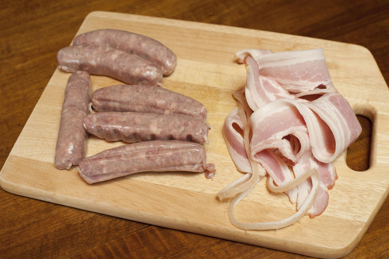 Making Pigs in Blankets with uncooked spicy sausages and rashers of bacon to form the outer wrapper ready on a wooden chopping board on a dark background