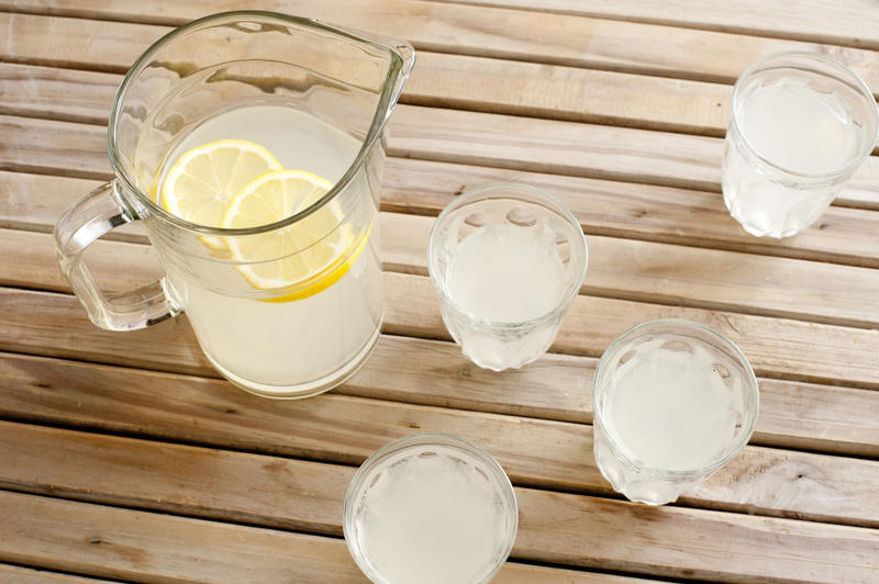 High Angle View of Pitcher of Freshly Squeezed Lemonade with Lemon Slices on Wooden Picnic Table Beside Four Glasses of Lemonade - Glasses of Refreshing Lemonade on Outdoor Table