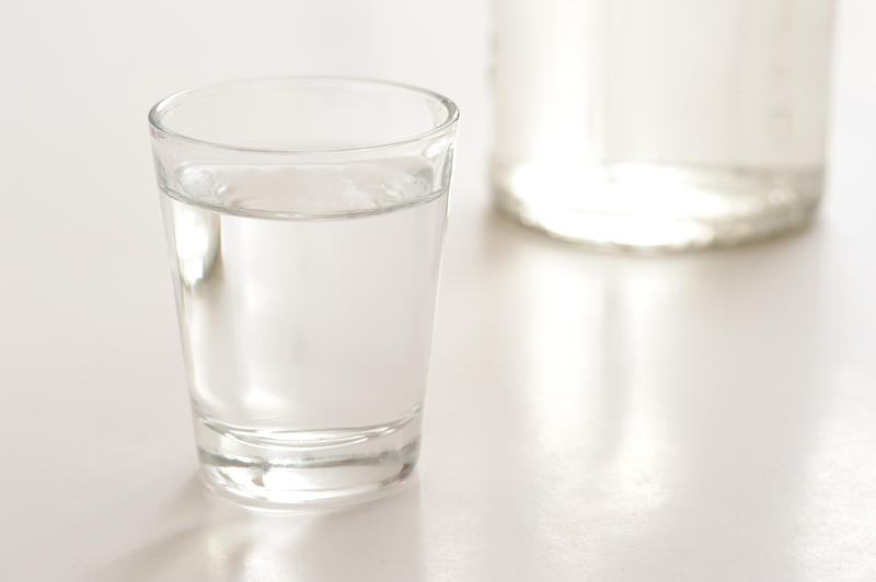 Shot glass filled with neat vodka standing on a reflective grey surface with the bottom of the bottle visible behind