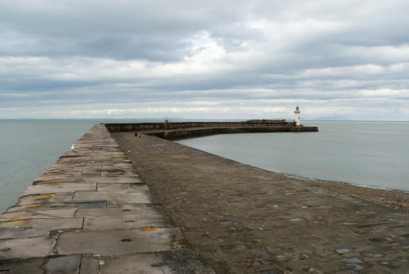 View along the deserted stone seawall at Whitehaven harbour to the small lighthouse and beacon at the end with a calm sea under clouds