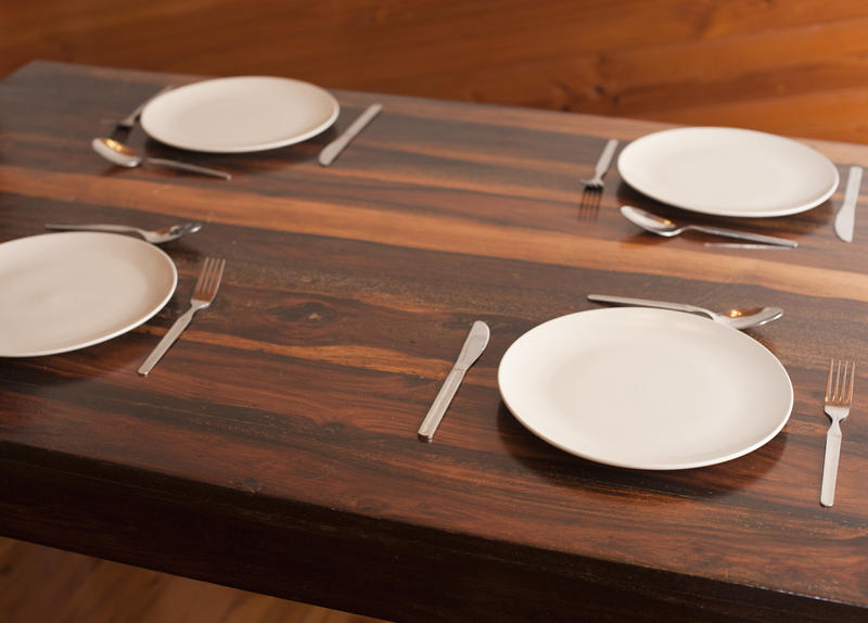 Four basic place settings on a wooden dinner table with plates and cutlery but no linen or placemats, closeup view