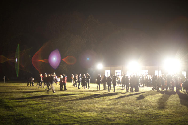 Crowd of people gathering on a sports field illuminated by bright spotlights as they prepare to celebrate Bonfire Night on 5th November